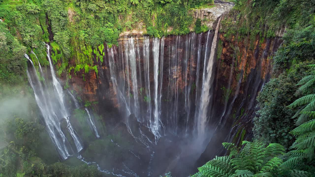 Video drone sudut tinggi yang merekam Air Terjun Tumpak Sewu di Indonesia dengan aliran air yang tak terhitung jumlahnya mengalir di atas dinding ngarai yang curam menciptakan kabut tebal, tanaman hijau subur, dan lanskap tropis yang kuat