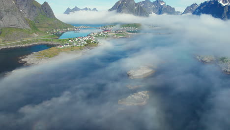 Lofoten-Islands,-Norway:-Aerial-shot-over-cloudy-mountains-and-small-islets-and-towards-the-fishing-town-of-Reine