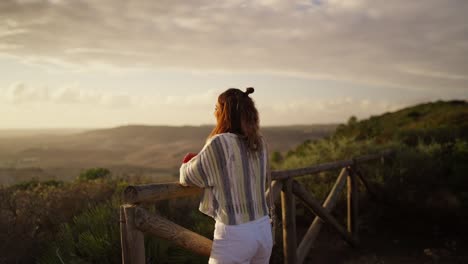 joven modelo morena apoyado en una barandilla de madera con vistas a las colinas al atardecer