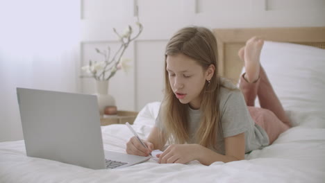 schoolgirl is chatting online by laptop from home lying on bed talking cheerfully and drawing in copybook