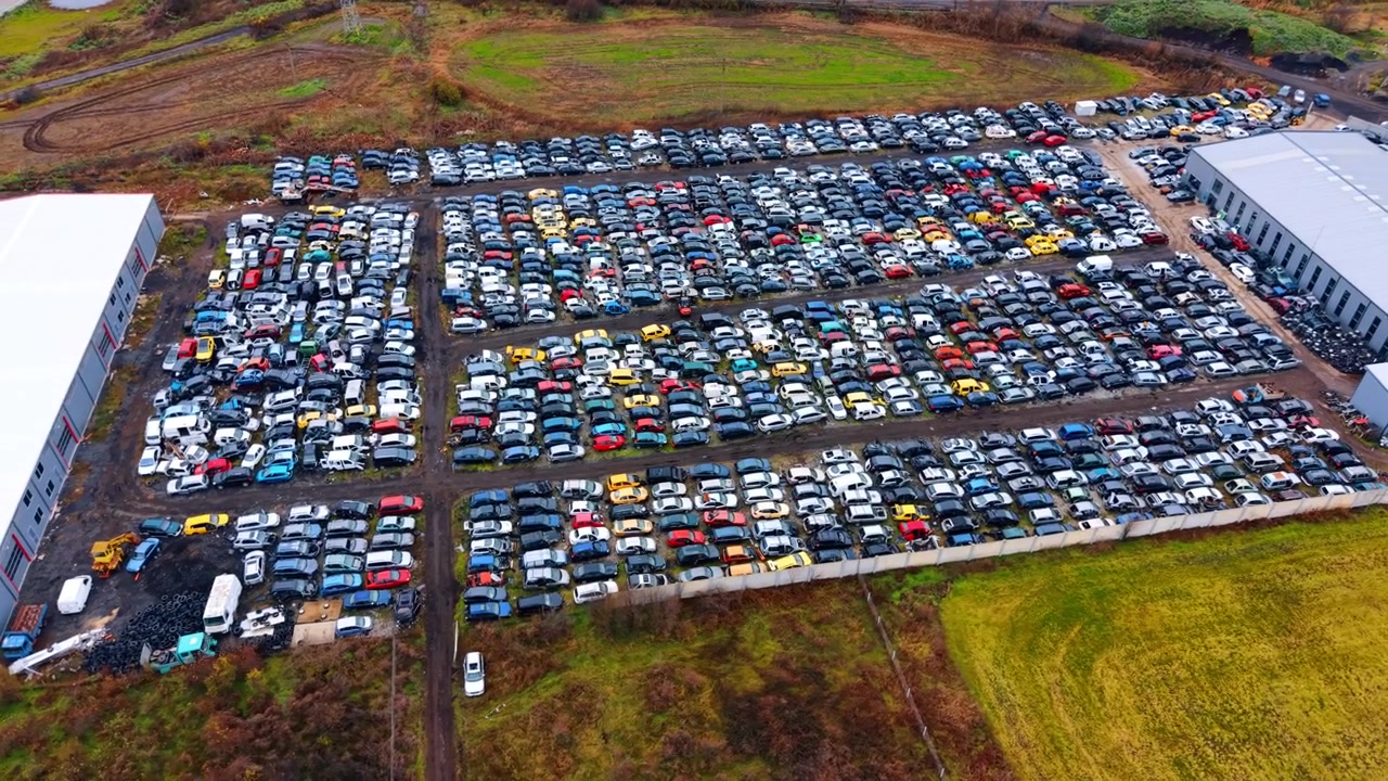 Bredt luftfoto af en massiv industriel bilophugningsplads. Panoramisk droneoptagelse af en enorm bilskrotplads ved siden af lagerbygninger og grønne marker under en overskyet himmel.