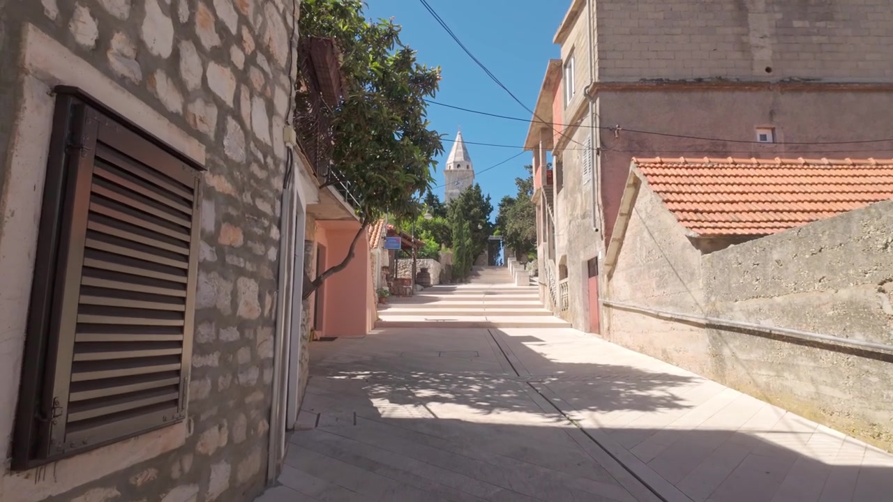 Bergauf gehen auf einer hellen Steinstraße in Primošten in Richtung des entfernten Kirchturms der St.-Georgs-Kirche, mit weißen Steinhäusern und einem sonnenbeschienenen Weg, der durch die Altstadt führt.