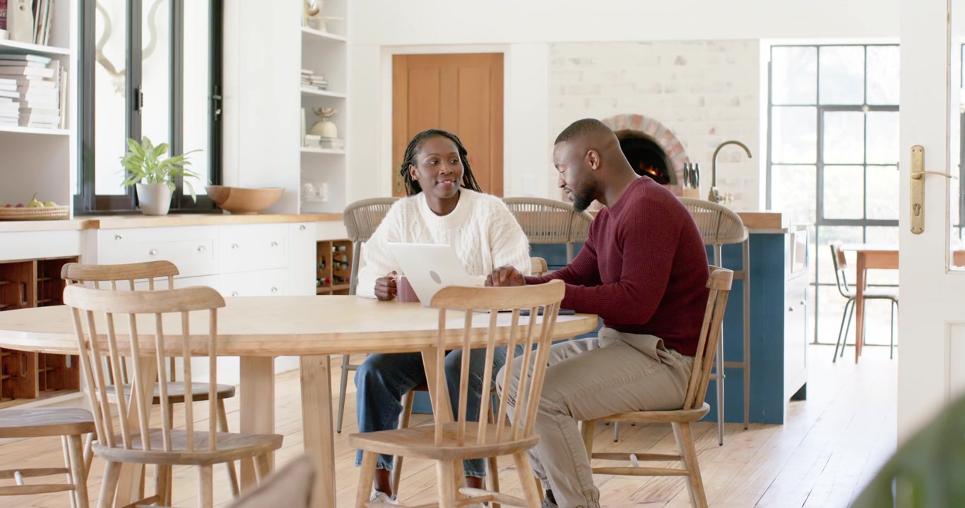 African couple at wooden table in kitchen woman showing white tablet man leaning tapping working