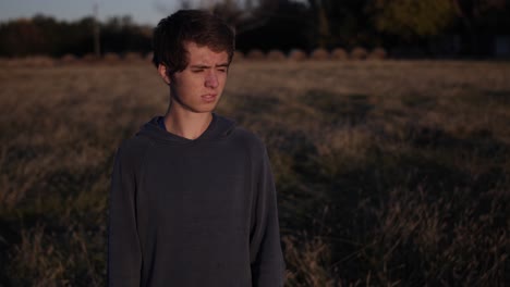 a-teenage-boy-stands-in-a-Kansas-field-looking-out-at-a-sunset-with-hay-bales-in-the-distance