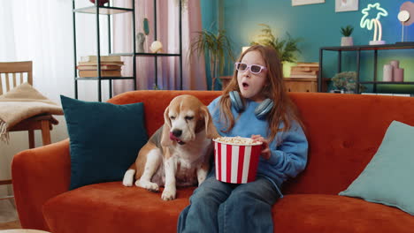 girl child in 3d glasses eating popcorn watching tv serial sport game film with beagle dog at home