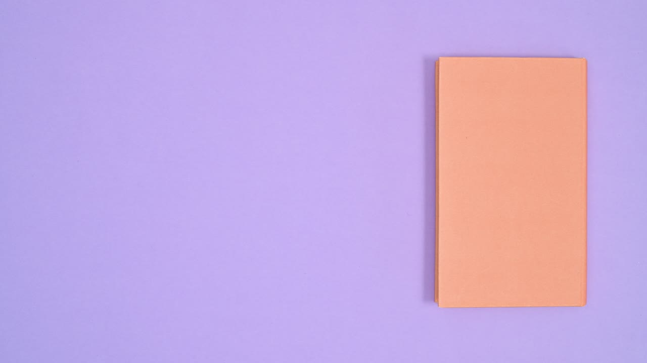 Orange hardcover vintage book move from right to left side of purple theme and open. Stop motion flat lay
