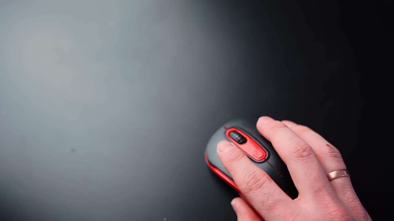 Top-down close-up of a hand resting on a black and red wireless mouse on a clean dark desk with large copy space, ideal for office productivity, UI navigation, and tech b-roll