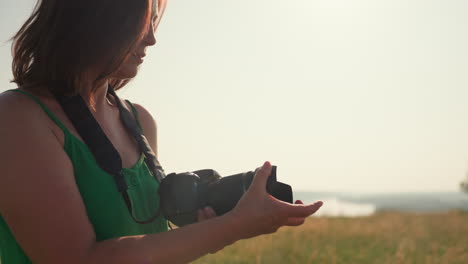 female photographer in green dress holding camera lowers hand to remove lens cover under bright sunlight in open field, standing in focus with serene face surrounded by golden grass and distant trees