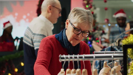 portrait of elderly woman browsing through clothes in christmas decorated clothing store during winter holiday season. aged customer at the mall in shopping spree session