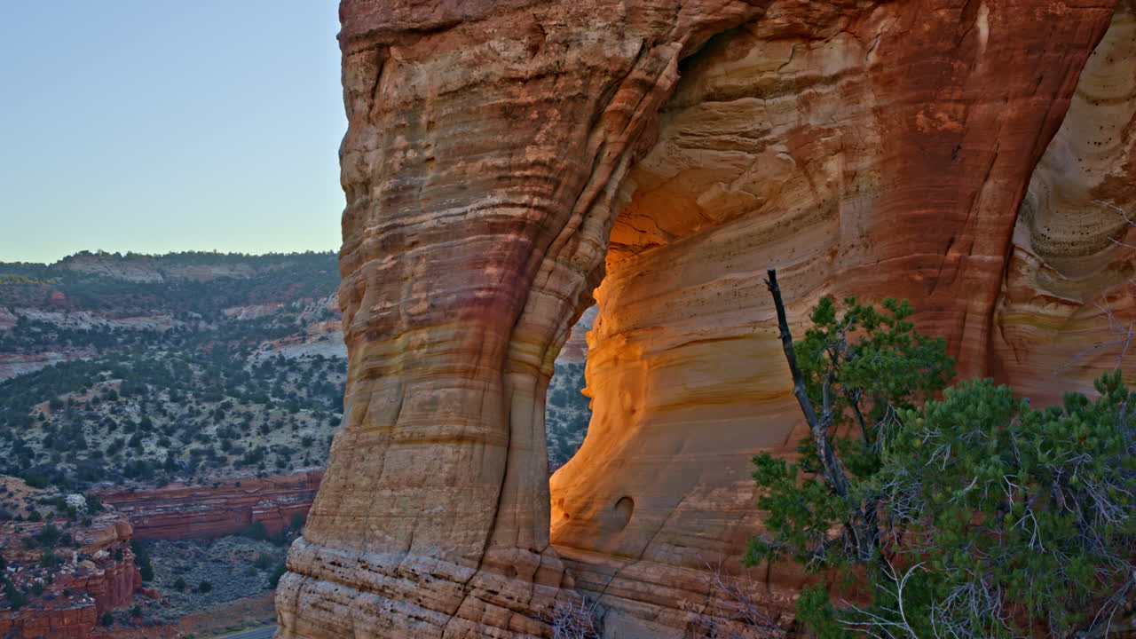 The drone gently flies toward a towering red rock arch, bathed in the soft, golden light of the rising sun.