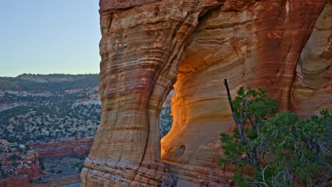 the drone gently flies toward a towering red rock arch, bathed in the soft, golden light of the rising sun.
