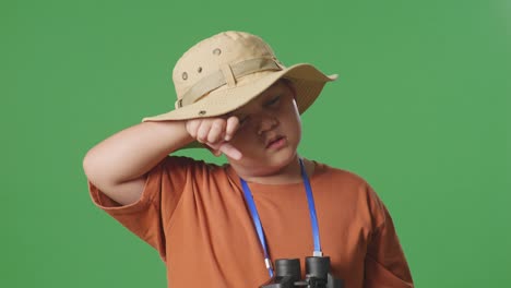 asian tourist boy with a hat wipes his sweaty face after looking through the binoculars. boy researcher examines something on the green screen background, travel tourism adventure concept, close up