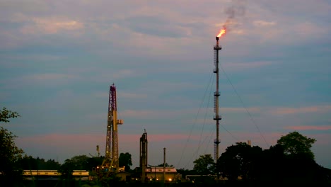 Fire-burning-on-top-of-gas-flare-stack-chimney-on-petroleum-refinery-against-sunset-red-sky