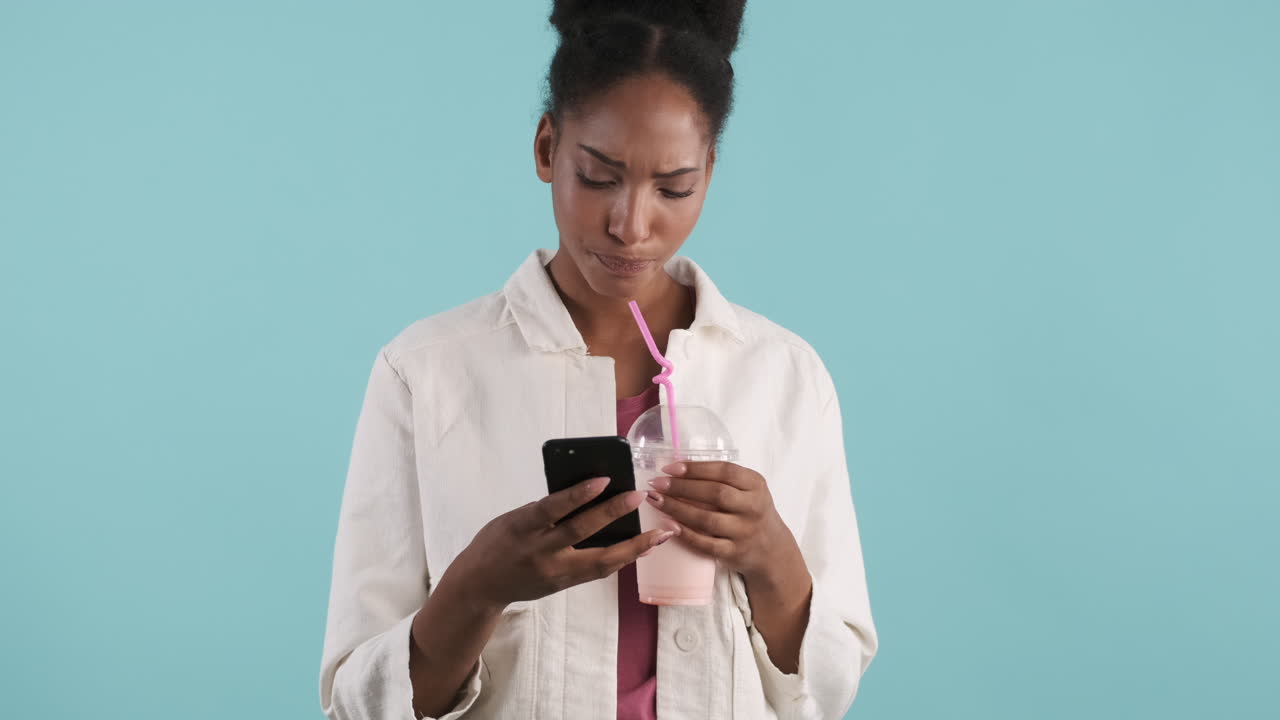 African american woman typing on phone while drinks milkshake