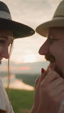 a wife in a white dress, a hat and bracelets, smiles lovingly as she touches her husband's face and beard in a grassy field at sunset. he wears a white shirt and a hat, and they share a joyful moment