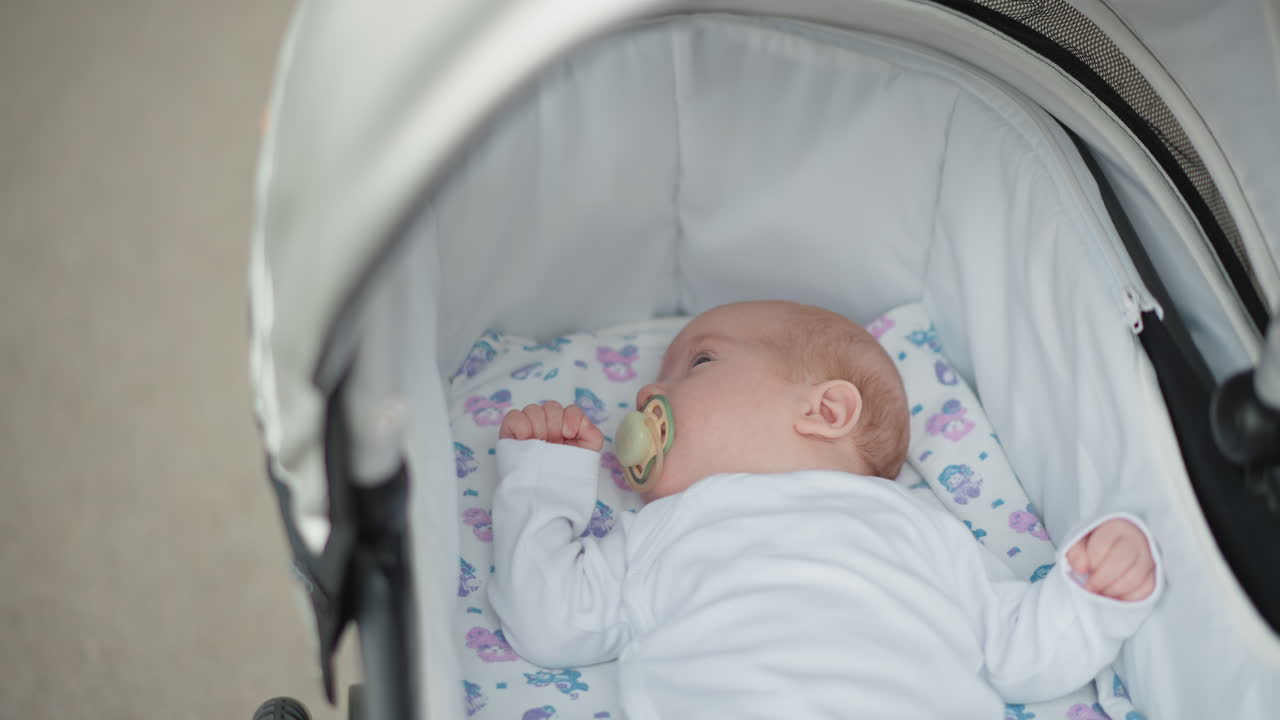 Neonato caucasico che dorme nel passeggino, con ciuccio su una coperta floreale, la cappottina soffice che incornicia il viso, illuminato da una delicata luce naturale. Con uno sfondo di marciapiede urbano, trasmette lo stile di vita e la dedizione dei genitori, ideale.
