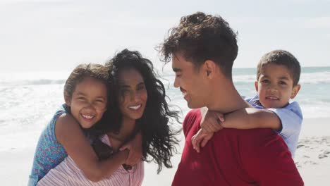 Portrait-of-happy-hispanic-parents-carrying-children-on-piggyback-on-beach