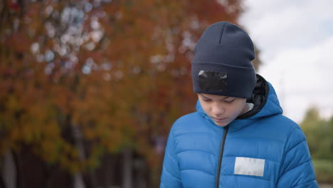 kid in blue jacket and black beanie walking with head bowed down thoughtfully, autumn trees in blurred background, reflective mood on a walk through the neighborhood