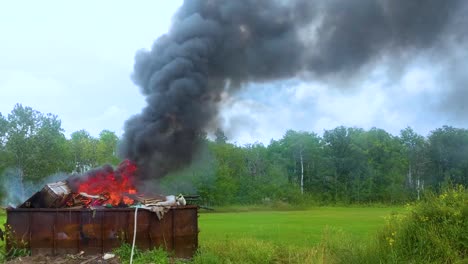 thick dark smoke billowing from a burning dumpster full of garbage on rural farmland