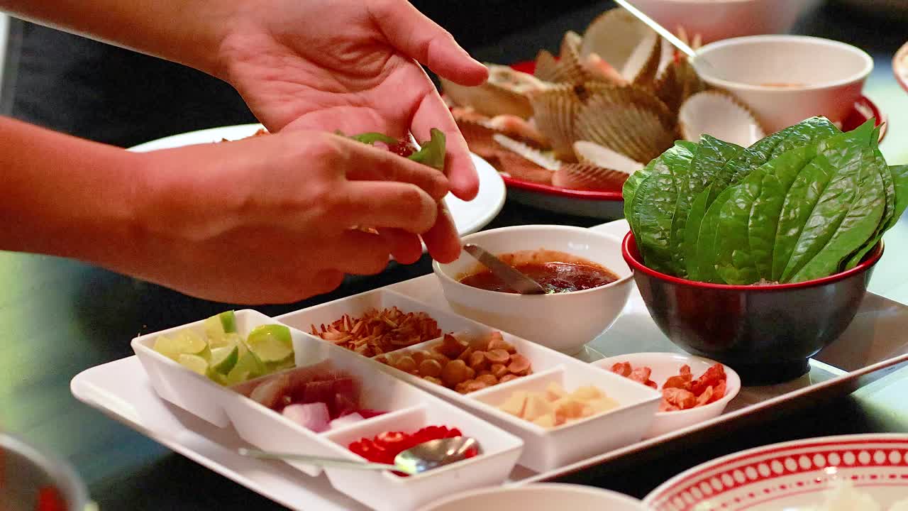 Hands assemble Miang Kham with fresh ingredients on a table in Phuket. Bright lighting highlights the vibrant colors and textures