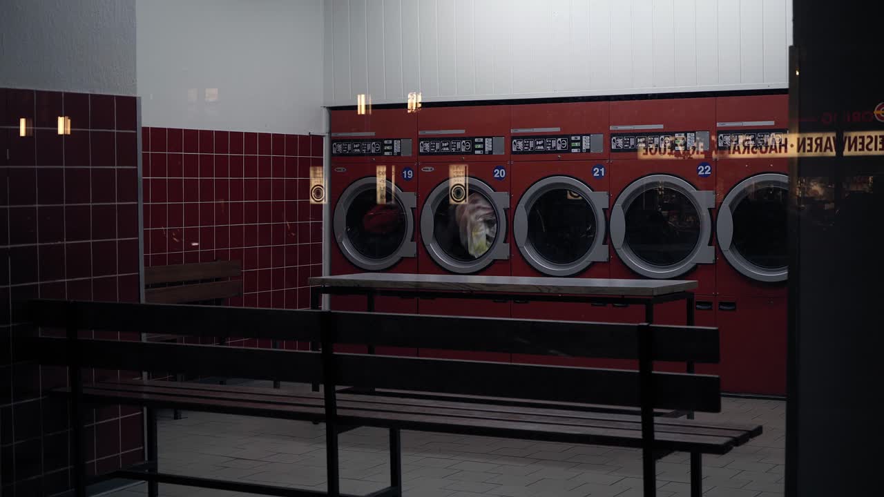 Shot of empty deserted self-service laundry shop at night, two laundry machines running, bench with no people in foreground