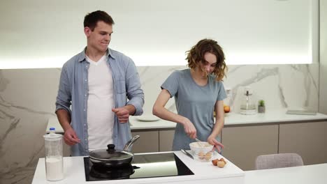 pareja alegre cocinando juntos en la cocina doméstica. mujer bonita positiva mezclando huevos para tortilla y el marido guapo está empezando a bailar. divirtiéndose juntos