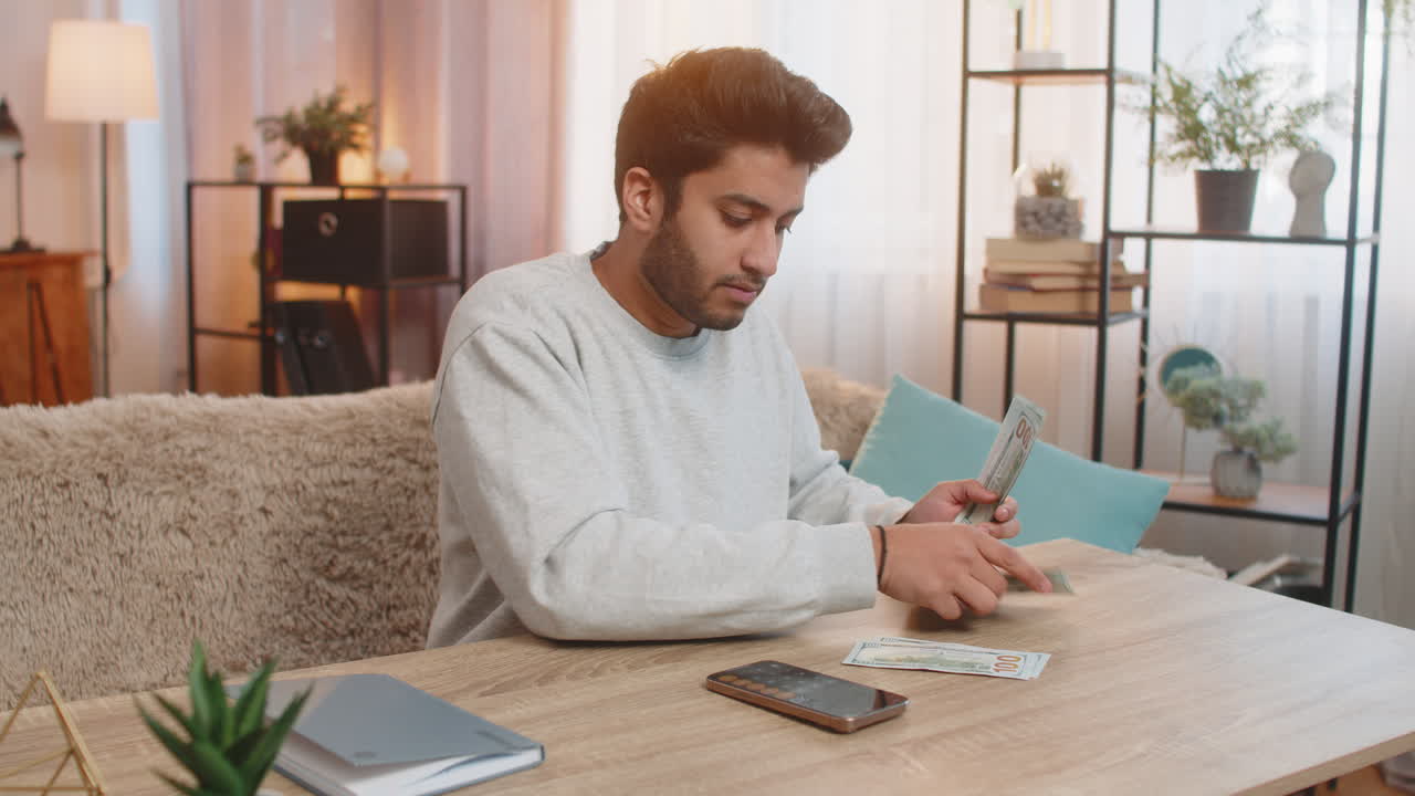 Young indian man planning expenses organizing cash in dollars using smartphone calculator at home