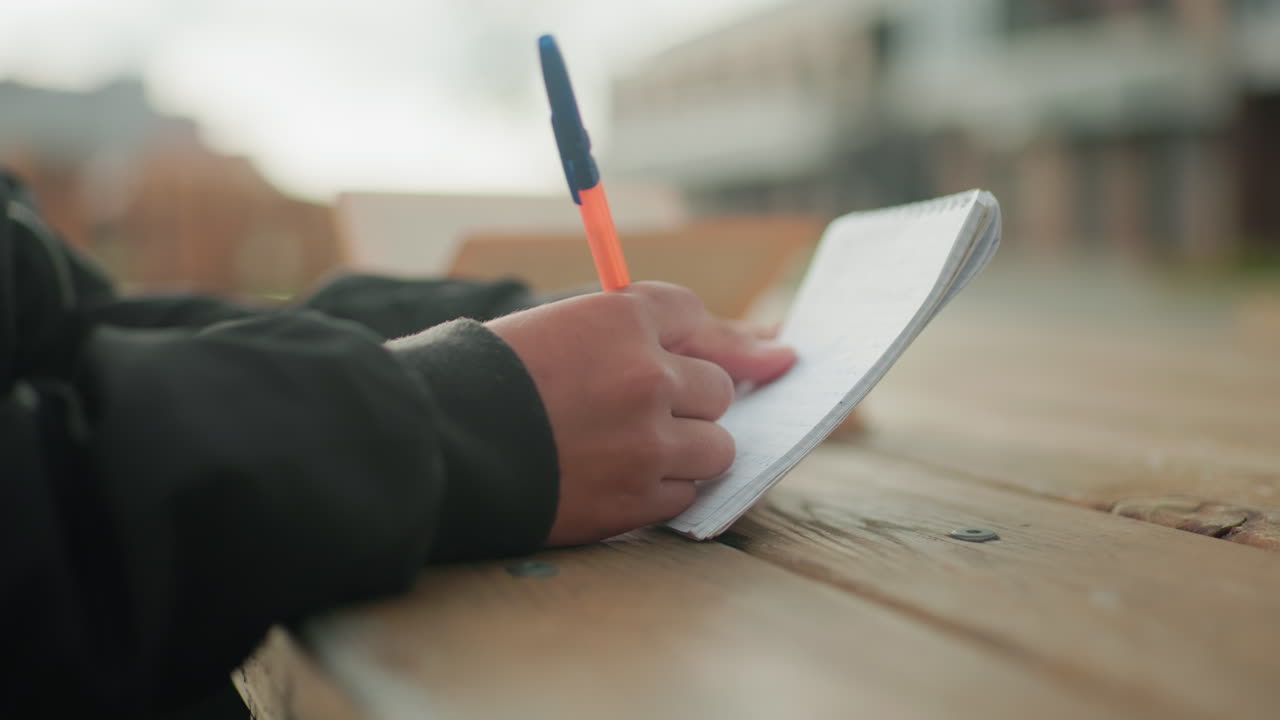 Close up side view of kid writing in notebook with pen on wooden table outdoors while blurred open book rests in background with pages flipping gently in wind