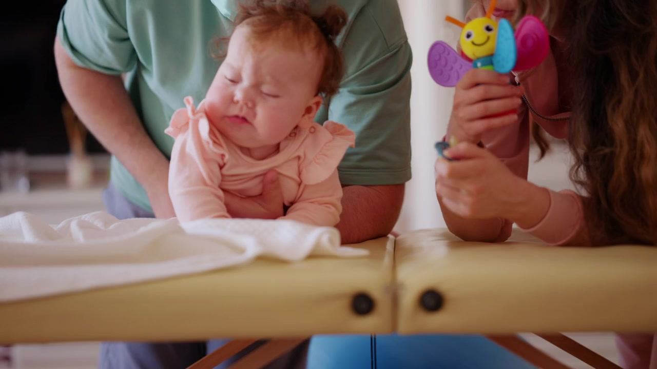 Um bebê deita-se confortavelmente enquanto observa um brinquedo colorido e faz exercícios de mobilidade com um cuidador.