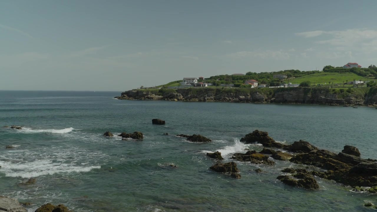 Smukt vidvinkelbillede af den barske kystlinje ved Playa del Rinconín i Gijón, Spanien. Med blide bølger, der bryder over klipper med frodige grønne beboelsesbakker i baggrunden under en klar himmel