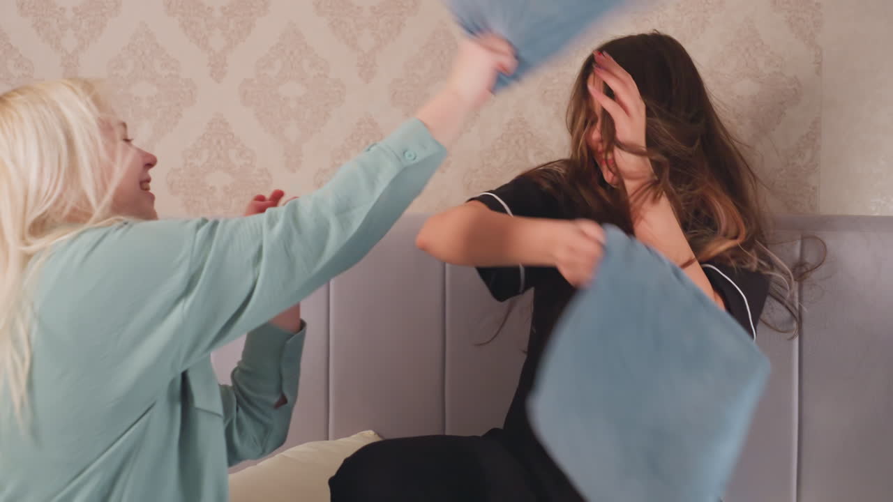 Close up of two siblings joyfully engaging in playful pillow fight on bed, both girls swing blue pillows at each other with laughter and energy, creating fun and cheerful atmosphere