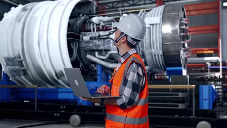 side view of asian male engineer with safety helmet working on a laptop and looking around while standing with airplane engine maintenance conducted, analytics checking the turbine
