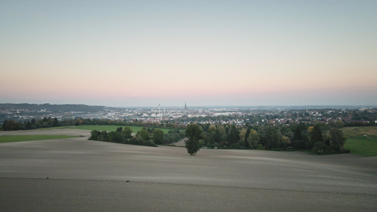 Scorrevole planata in avanti con drone 4K sopra cime di colline erbose verso lo skyline di Ulm al tramonto, con campi ondulati e un orizzonte dai colori pastello.