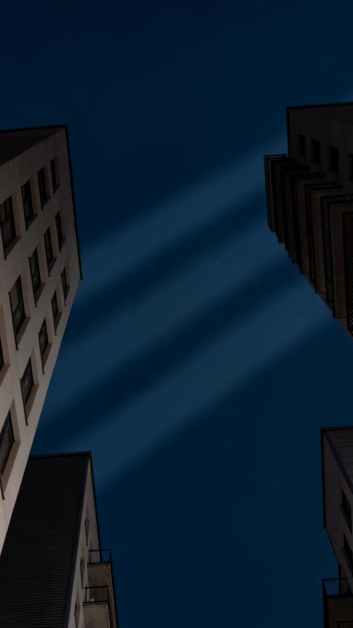 Video vertical: Tres haces de luz comienzan a barrer el cielo nocturno sobre un cañón urbano, iluminando ventanas.