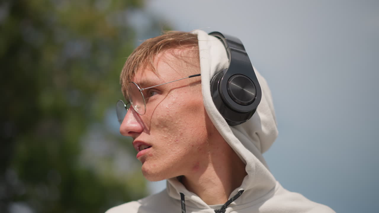 Uomo caucasico con cappuccio e cuffie che ascolta attentamente, esercitandosi nel fraseggio vocale e alla ricerca di una melodia, primi piani espressivi con cielo nuvoloso, con uno squarcio di sole e un dettaglio sugli occhiali