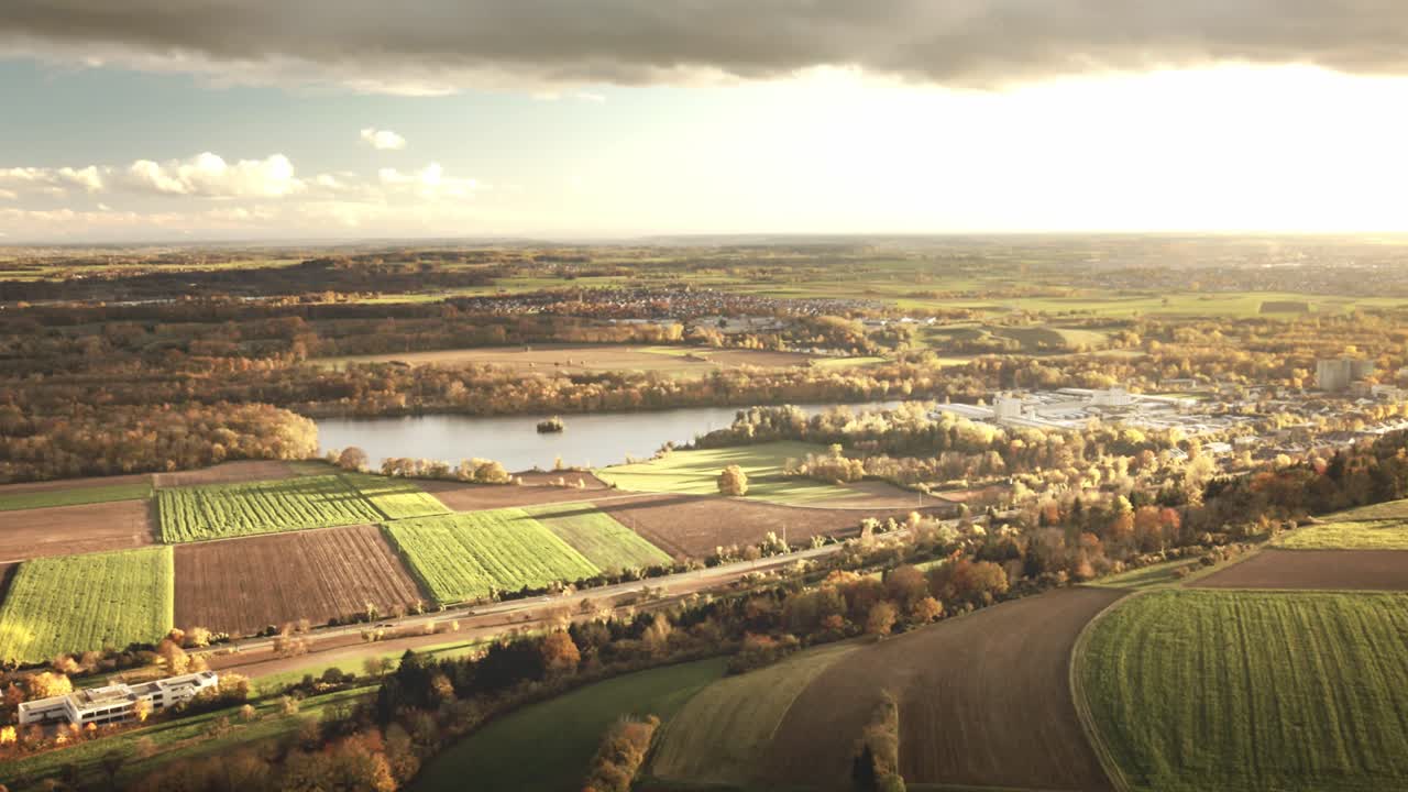 Camera glides forward above rural fields glowing in evening light, unveiling a winding lake, distant forests, and scattered villages under dramatic clouds