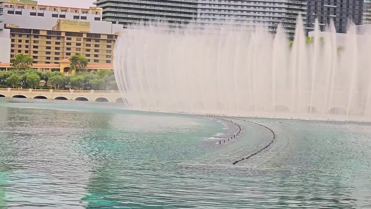 Espectáculo escénico de las Fuentes del Bellagio en Las Vegas, con chorros de agua curvos, niebla, lago azul verdoso, edificios de resorts, palmeras, hoteles de gran altura y el famoso horizonte del Strip.
