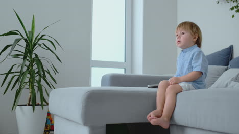 a 2 years old boy sits on a sofa and watches tv sitting with a remote control in his hands.