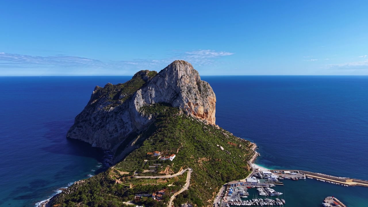 Ikonická skalní formace Penon de Ifach se tyčí nad Středozemním mořem v Calpe ve Španělsku, s viditelným přístavem a bujnou zelenou vegetací u jejího úpatí.