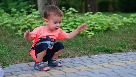 adorable caucasian toddler sits squatting outdoors. baby is watching the insect running by the ground.