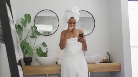 happy african american woman brushing teeth and using smartphone in bathroom