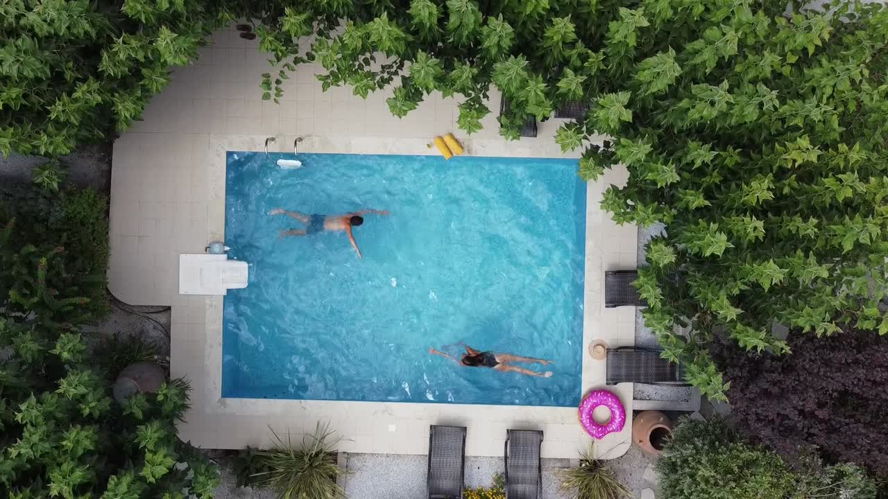 Young couple enjoying summer by pool - both swimming in pool - drone hovering over pool in bird's eye view