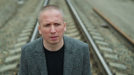 a close view of an aged white man wearing a grey suit and a black polo shirt, walking on a railway with stones, with view blur of trees and electric poles on either side