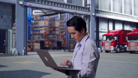 side view of an asian male professional worker standing with his laptop, outside of logistics distributions warehouse, typing on his laptop's keybaord with meditation