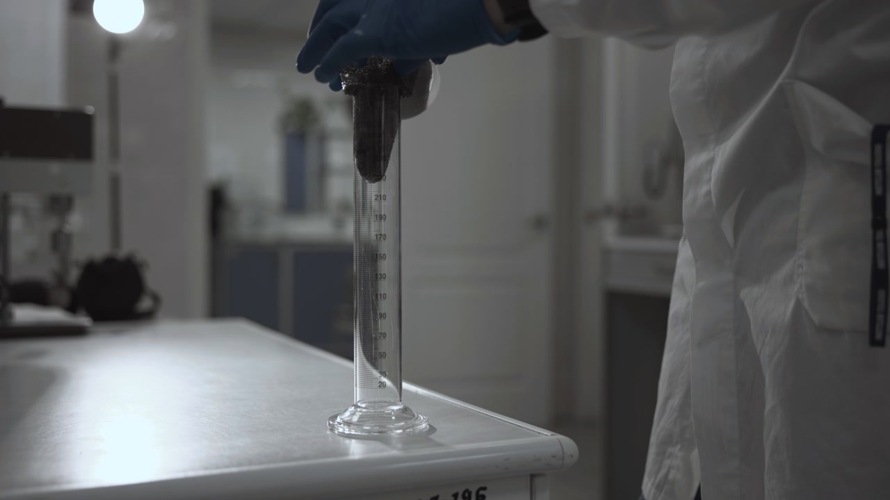 Técnico con guantes y bata blanca de laboratorio dispensa cuidadosamente una muestra de líquido oscuro en una probeta graduada alta y transparente.