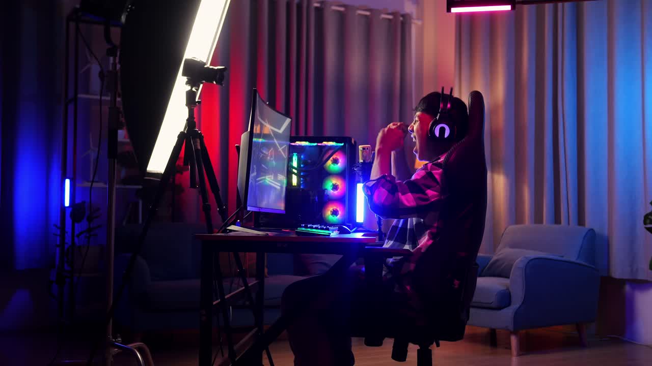 vista lateral de un niño asiático que grita gol y baila celebrando el juego ganador a través de la red en una computadora personal. videojuego de transmisión en vivo, escritorio iluminado por luz de tira led rgb