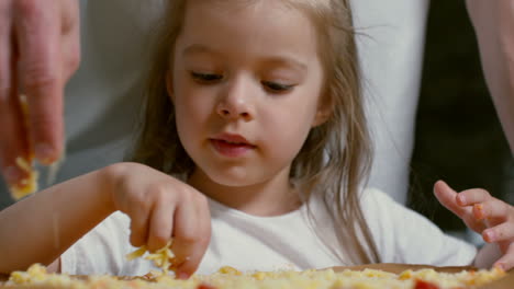 Close-Up-View-Of-Cute-Little-Girl-Of-Primary-School-Age-Sprinkling-Grated-Cheese-On-Pizza-When-Cooking-Together-With-Father