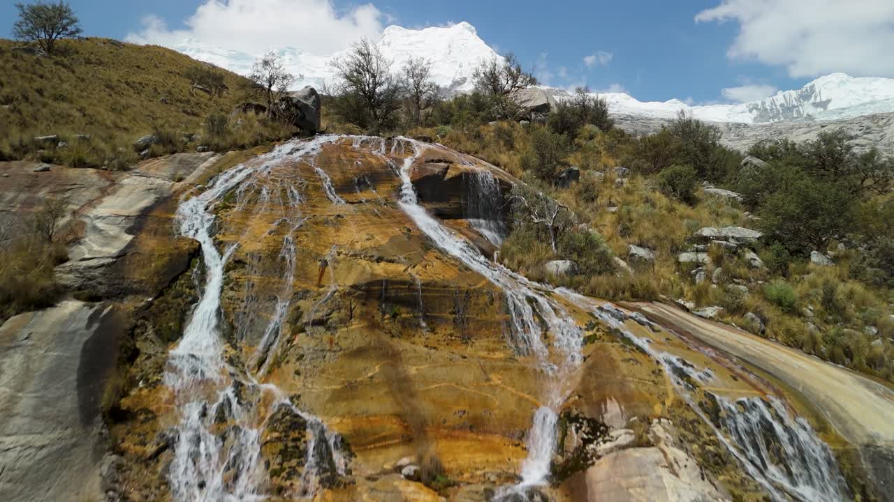 A spectacular aerial view rises above a beautiful mountain waterfall, revealing the majestic, snow-capped peaks of the Andes towering in the background near Laguna 513 in Peru