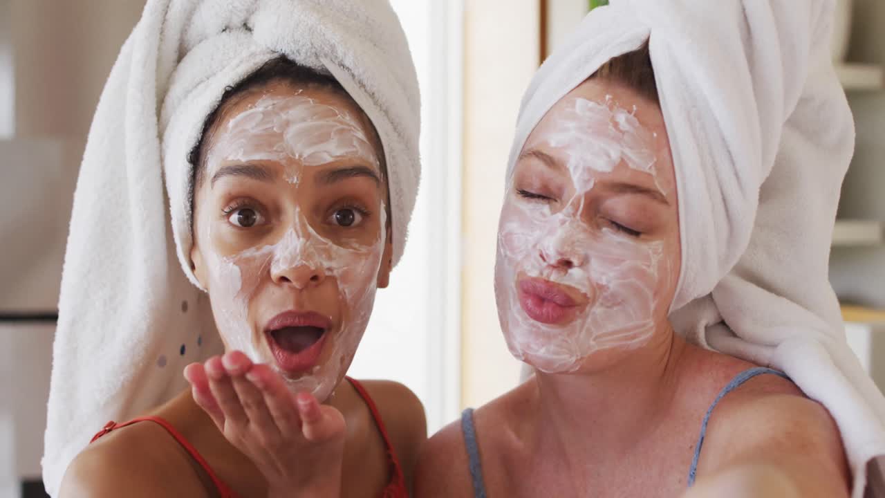 Diverse happy female friends wearing towels on heads and cleansing masks taking selfie at home