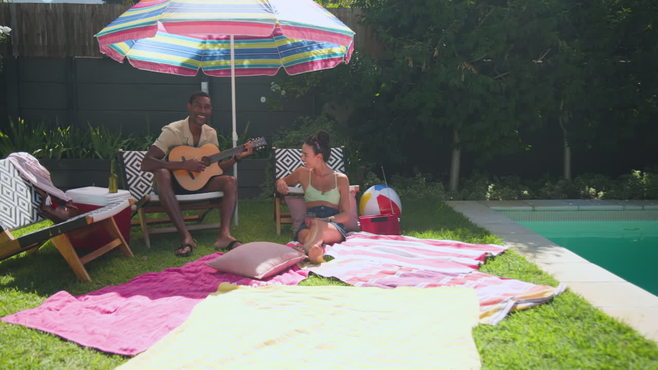 Diverse friends relaxing by pool, man playing guitar, woman laughing under colorful umbrella